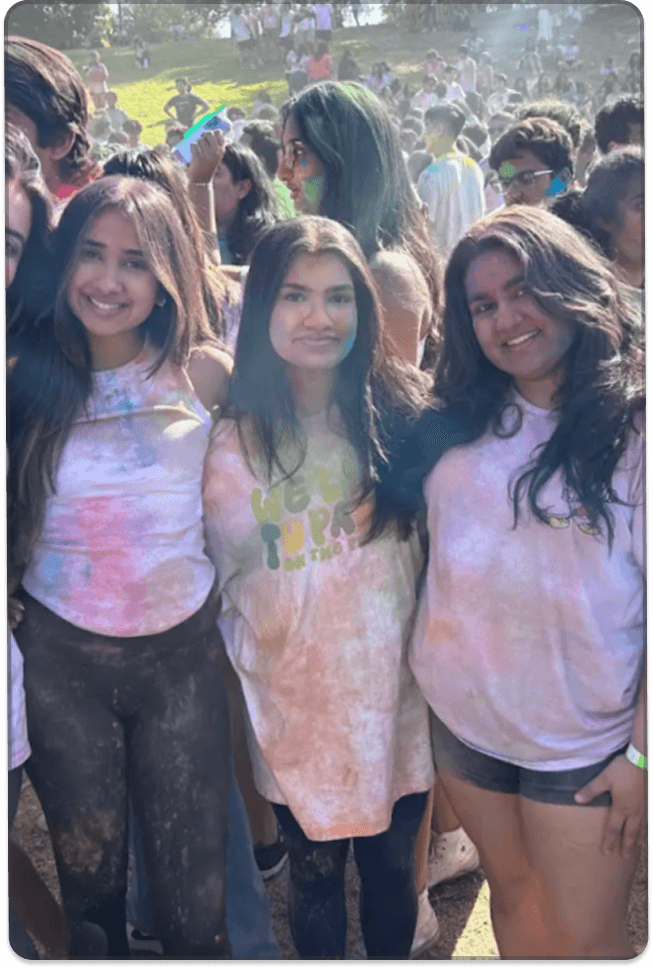 A group of young people pose together in a crowded outdoor setting, with colorful powder visible on their clothes and faces.