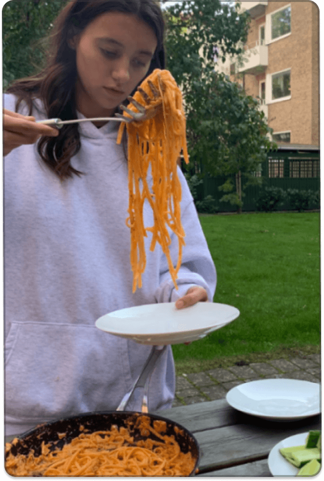 A young person holds a plate of noodles while standing beside a table with food, including a pan of noodles and sliced vegetables, in a residential outdoor area.