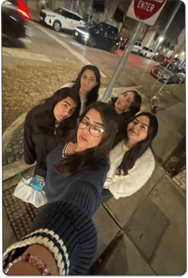 A group of young people pose together on a sidewalk at night, smiling toward the camera, with parked cars and street signs in the background.