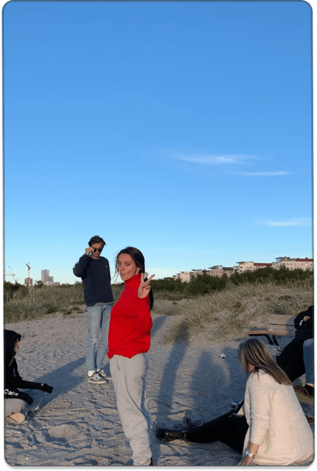 A group of young people spend time together on a sandy beach or dune area under a clear blue sky, with several people standing and sitting in the background.